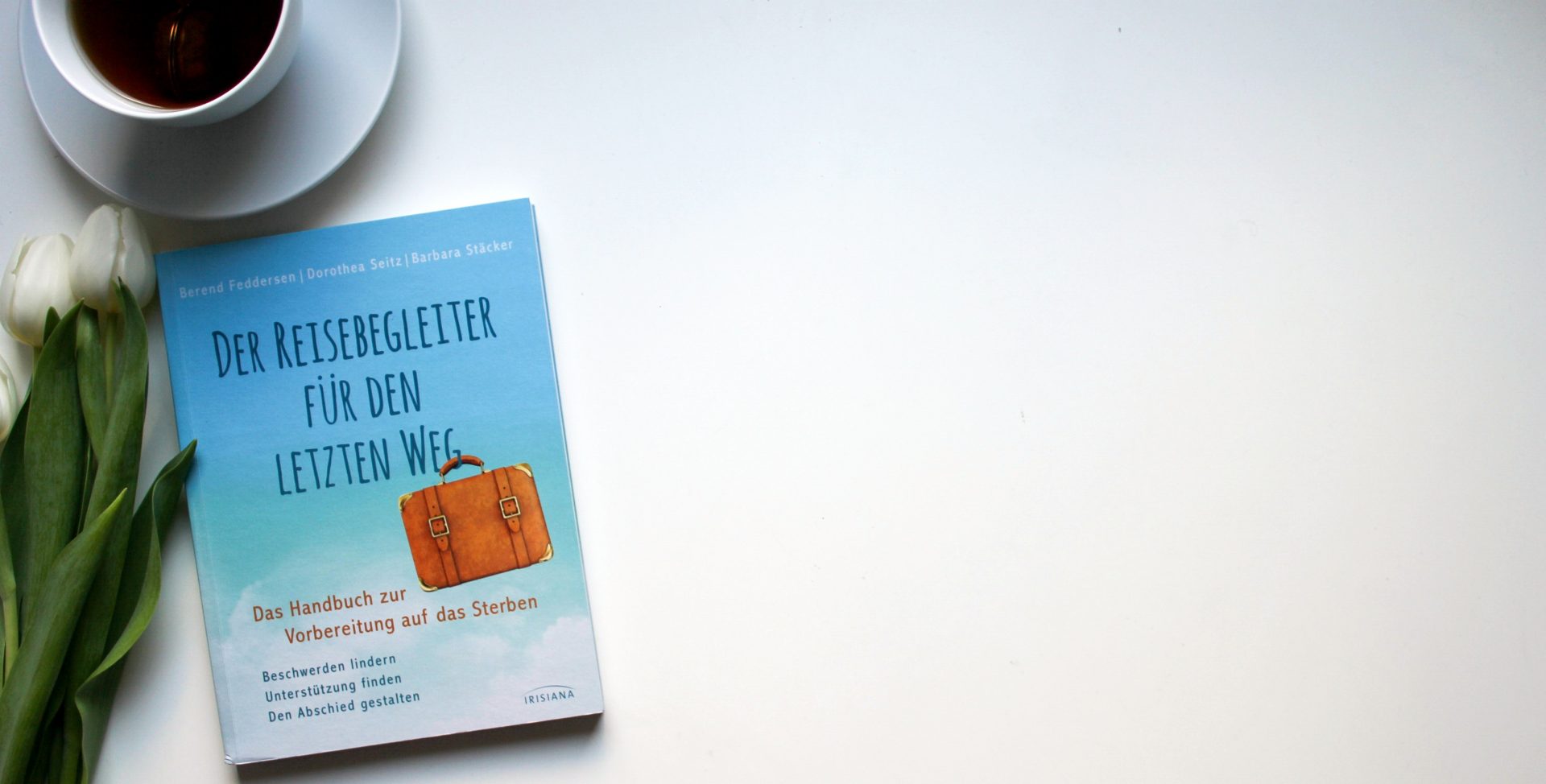 Buch liegt auf einem weißen Tisch neben einer Tasse Tee und weißen Blumen