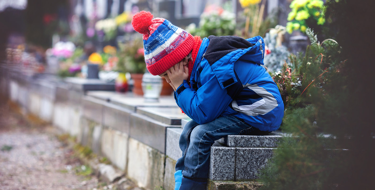 Trauriger kleiner Junge, der auf einem Grab auf einem Friedhof sitzt