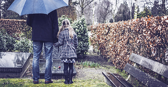 Vater steht mit Tochter mit einem Regenschirm vor einem Grab