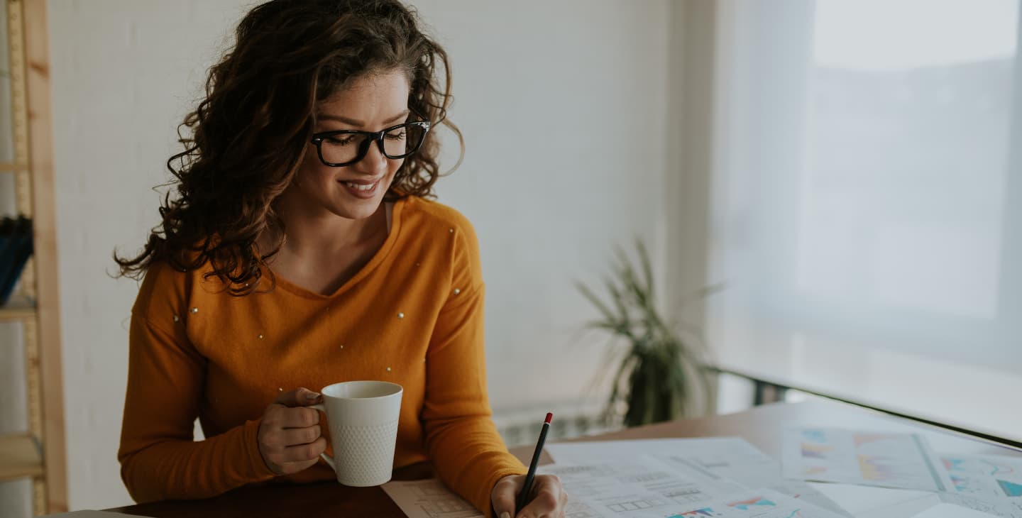 Frau mit Brille hält eine Tasse und schreibt mit Stift auf Arbeitsunterlagen