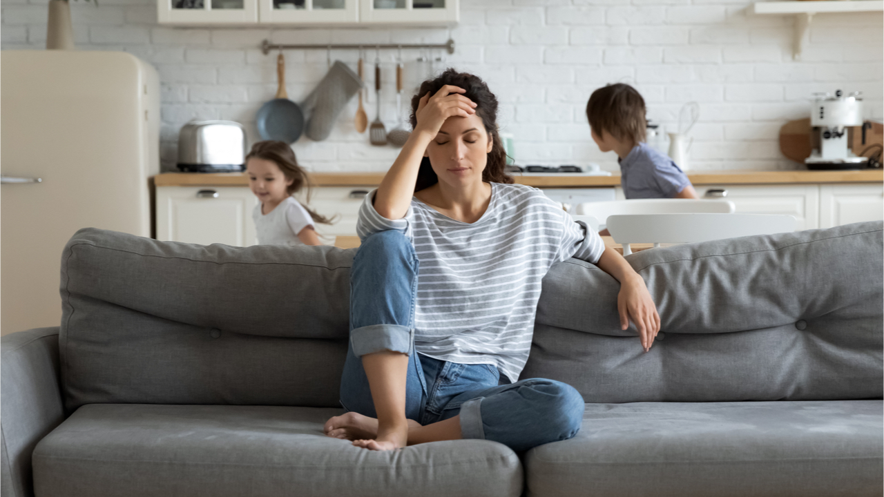 Frau sitzt gestresst auf dem Sofa und im Hintergrund spielen Kinder